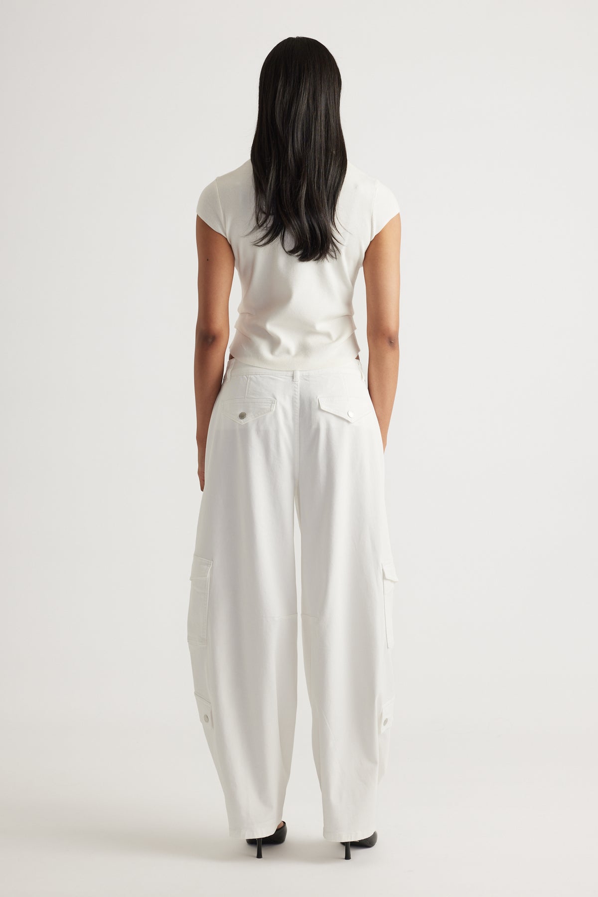 Rear view of a woman wearing the Lily Pleat Detail Knit Tee in soft winter white paired with wide-leg white pants by DECJUBA