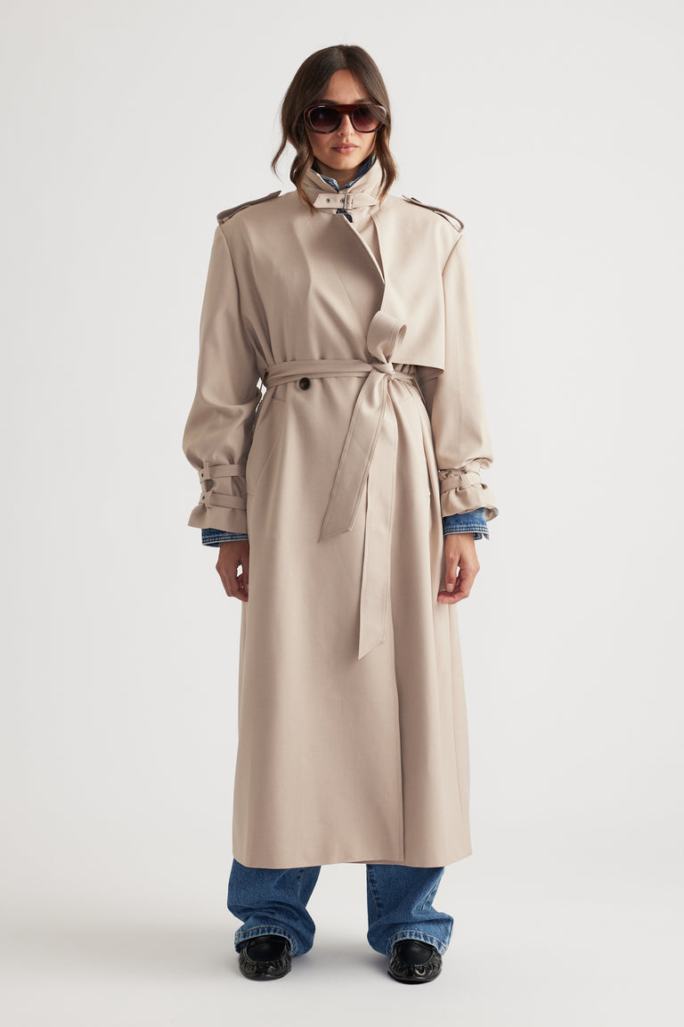 Woman wearing a classic beige trench coat with a belted waist, paired with blue jeans and black loafers, accessorized with sunglasses
