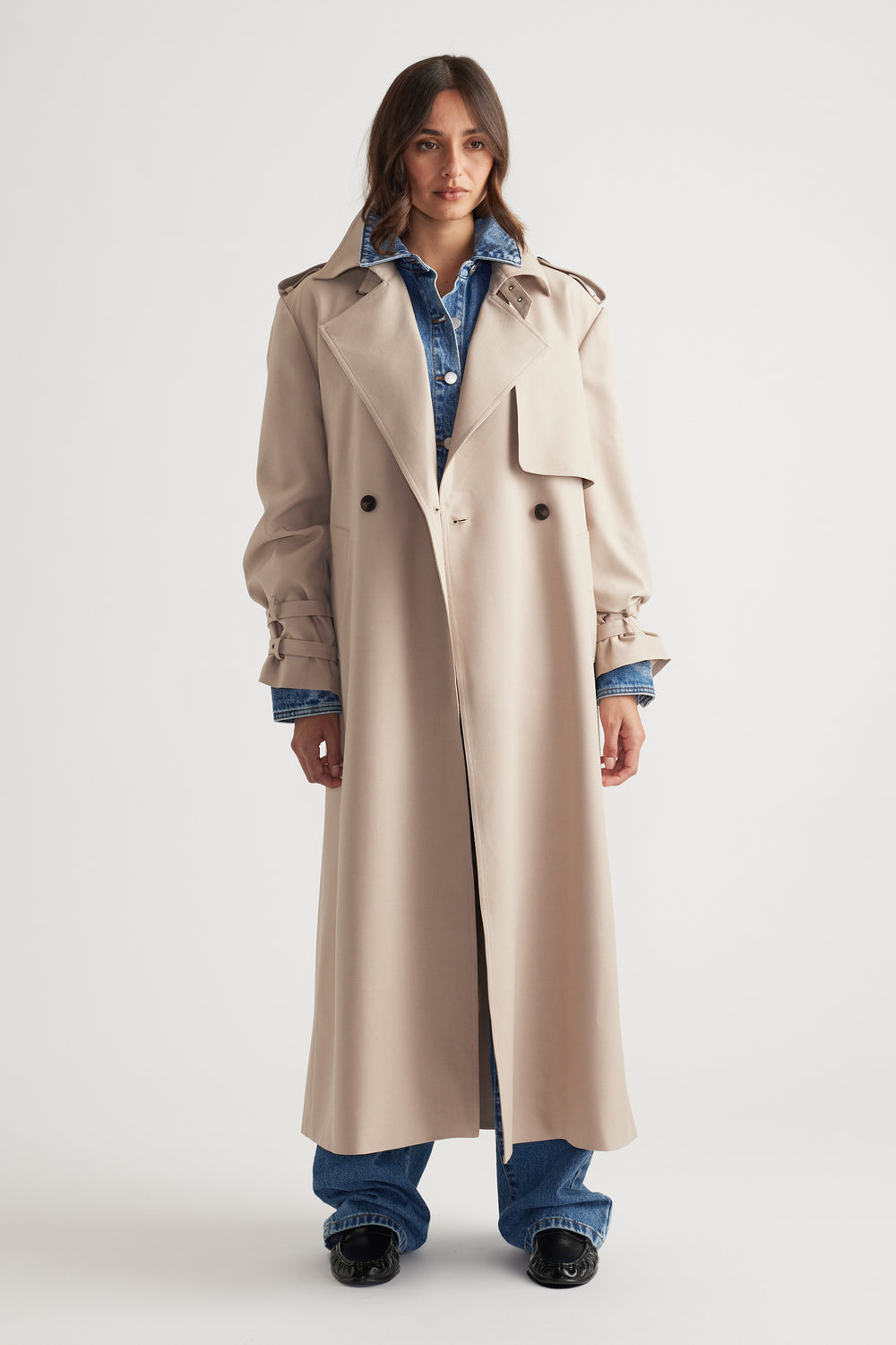 Woman wearing a long beige trench coat with gathered cuffs layered over a blue denim jacket and matching jeans