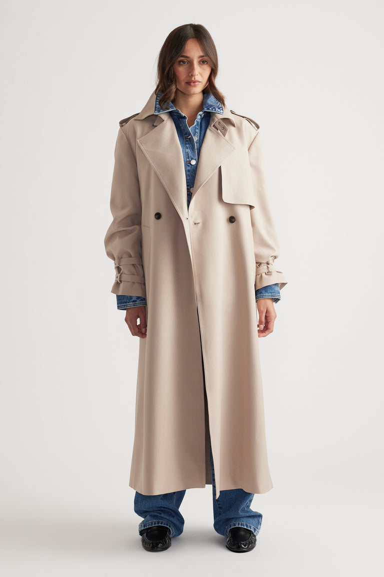 Woman wearing a long beige trench coat with gathered cuffs layered over a blue denim jacket and matching jeans