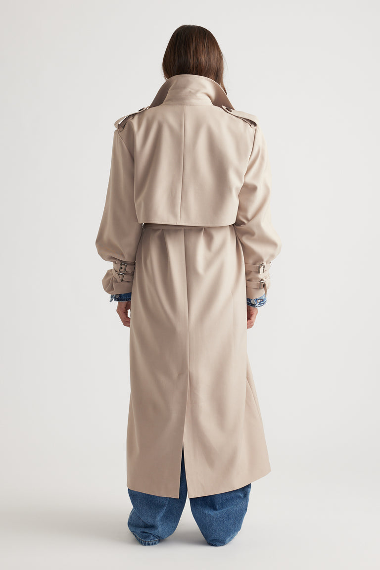 Woman wearing a biscuit-colored long trench coat with belted cuffs and back yoke, paired with loose blue denim jeans