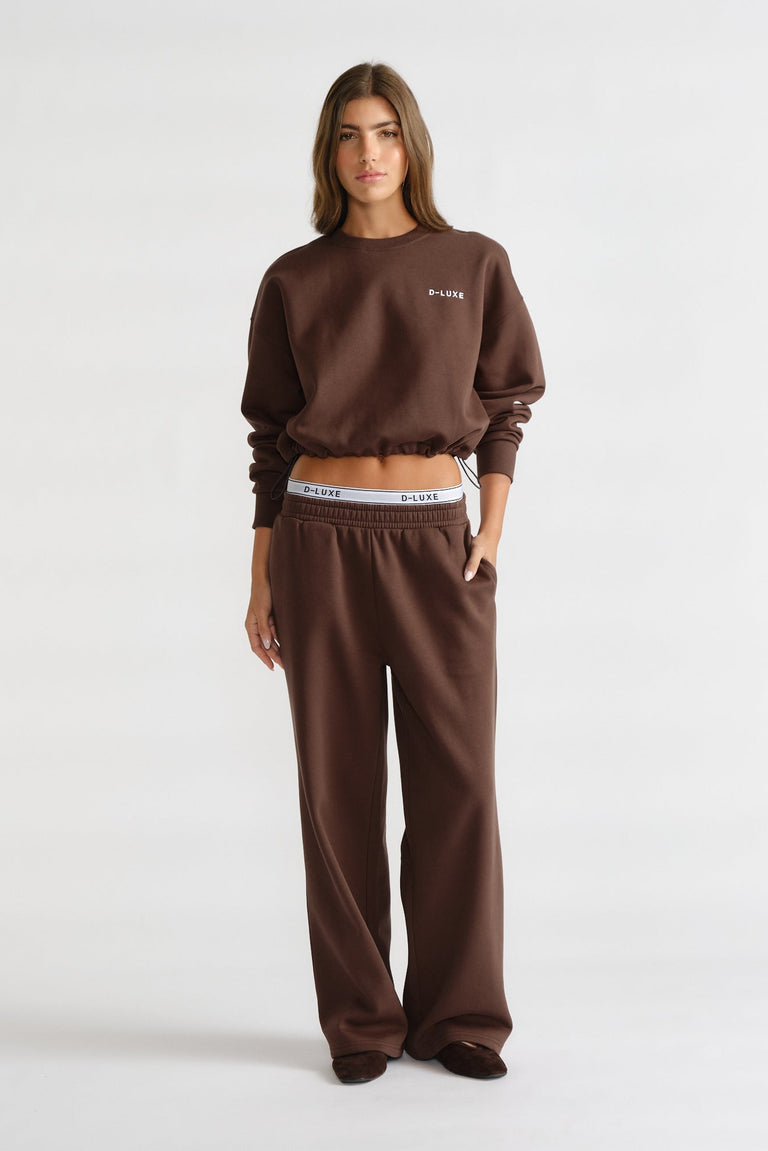 Woman wearing a relaxed dark brown sweat set with elasticized waistband trackpants and a cropped sweatshirt with minimal logo detail
