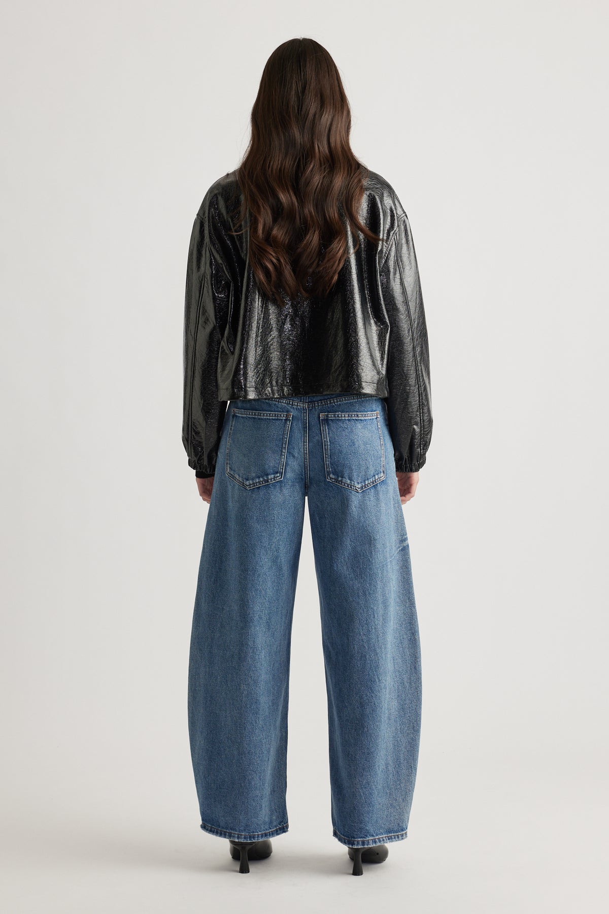 Woman wearing a cropped black shiny zip jacket paired with relaxed wide-leg blue denim jeans and black heels