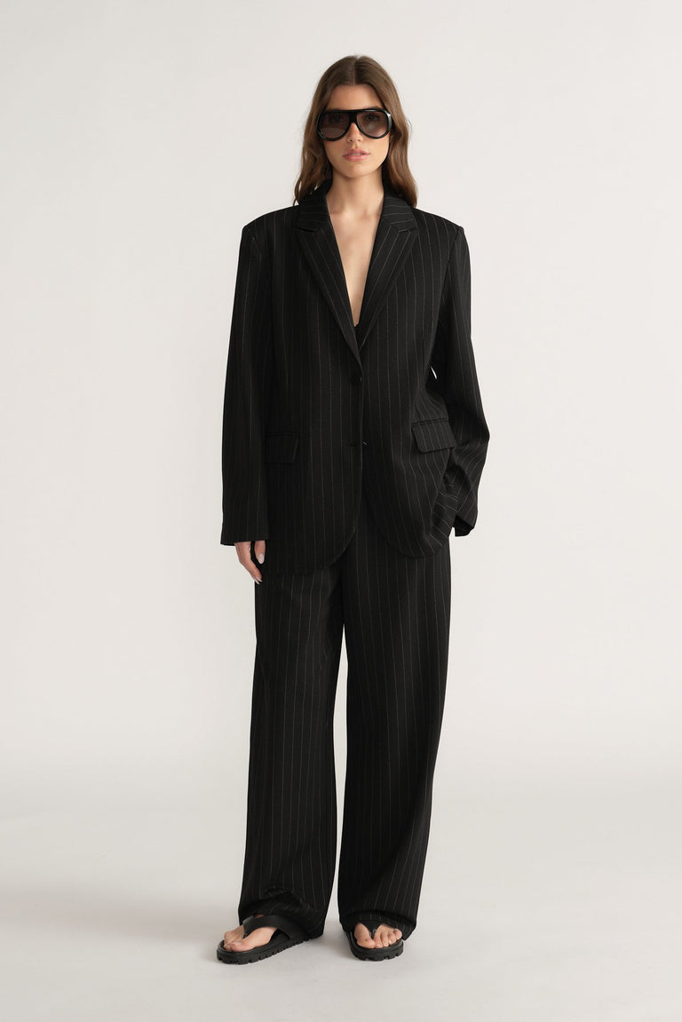 Woman wearing relaxed fit black pinstripe blazer and matching wide-leg pants with oversized sunglasses on plain background