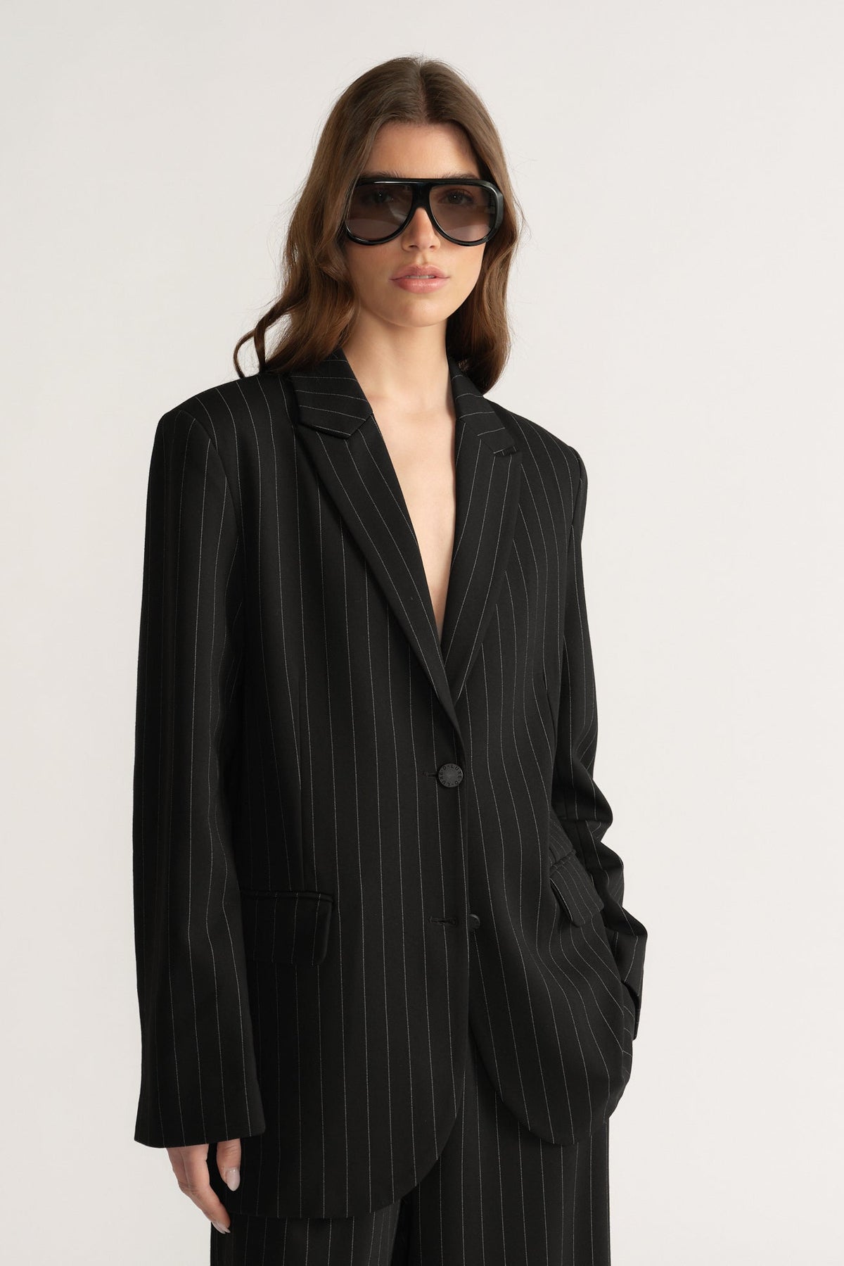 Woman wearing oversized black pinstripe blazer with notch lapels and welt pockets, styled with dark sunglasses