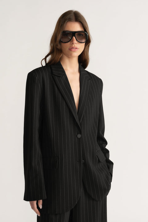 Woman wearing oversized black pinstripe blazer with notch lapels and welt pockets, styled with dark sunglasses