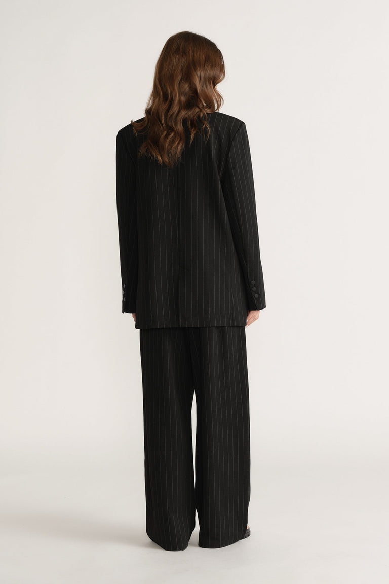 Woman wearing black pinstripe blazer and wide-leg pants, shown from the back in a minimalist studio setting