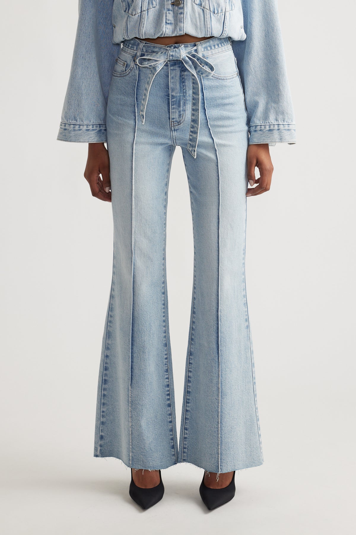 Woman wearing light blue Freya Flare Jean with a tied waist belt, styled with matching denim jacket and black heels