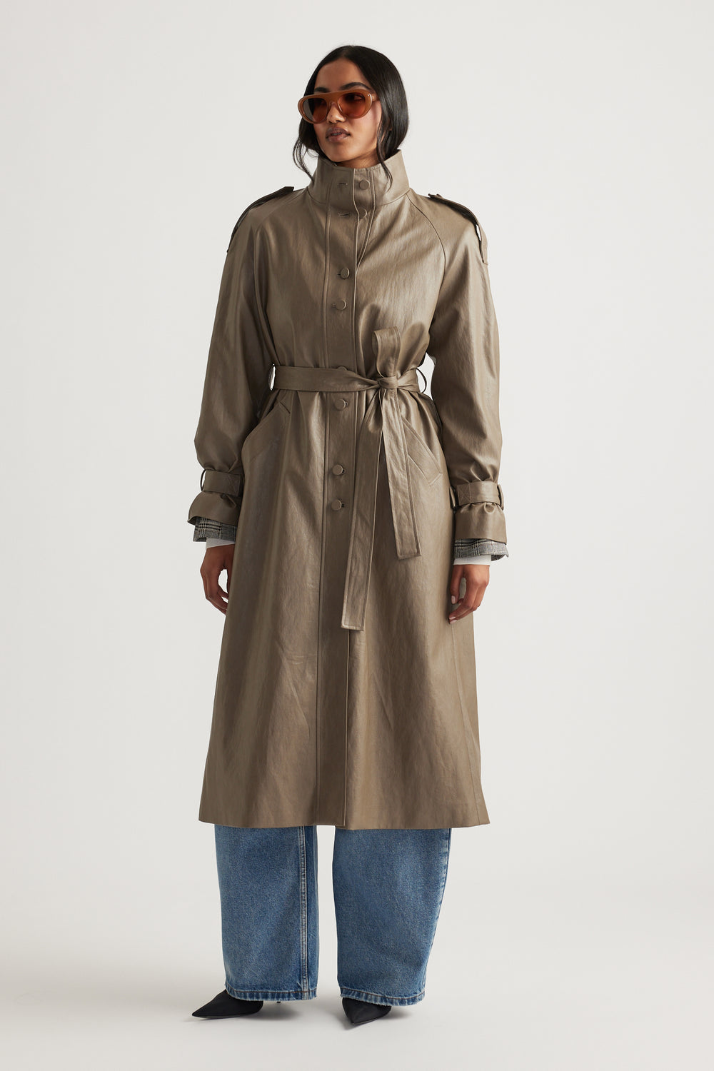 Woman wearing a taupe faux leather trench coat with a belted waist, styled with wide-leg denim jeans and sunglasses