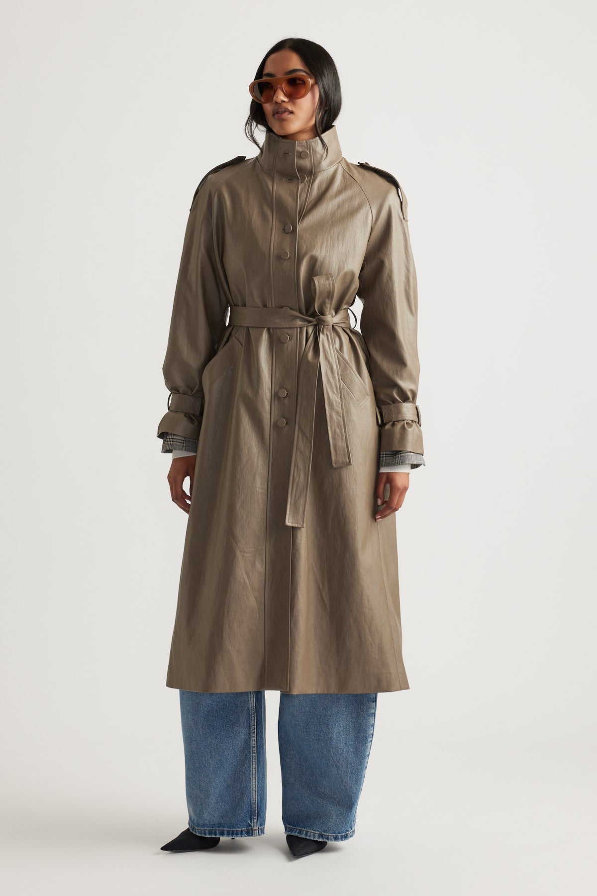 Woman wearing a taupe faux leather trench coat with a belted waist, styled with wide-leg denim jeans and sunglasses