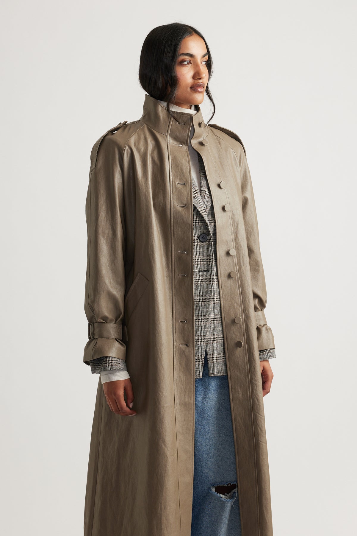 Woman wearing a taupe faux leather trench coat layered over a plaid blazer and distressed blue jeans for a chic look