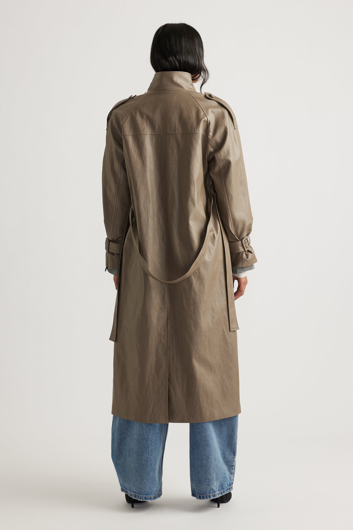 Back view of a taupe faux leather trench coat with belt and cuff straps, paired with relaxed blue jeans and black heels.