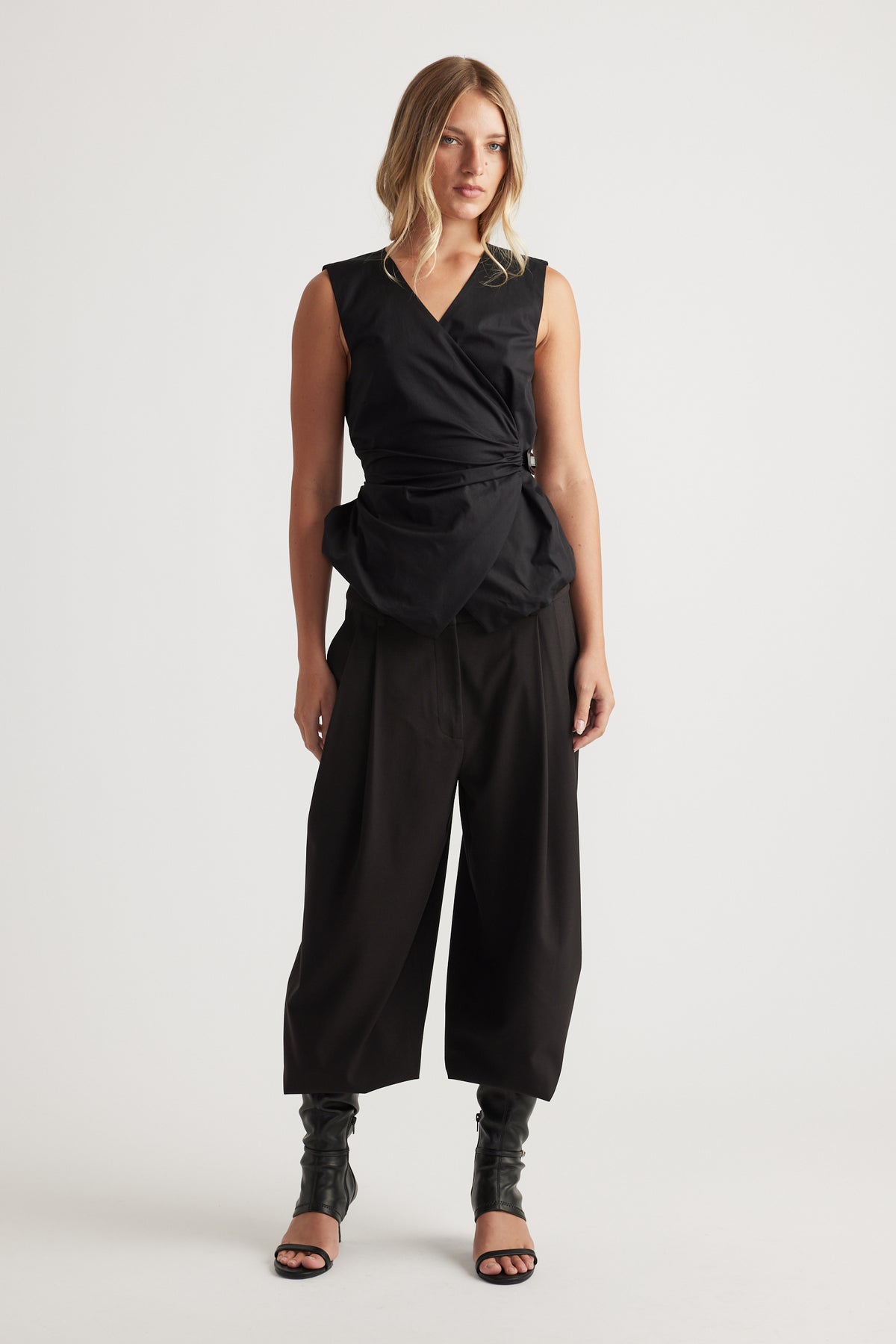 Woman wearing Ava Belted Wrap Top in black with sleeveless cut and adjustable side belt, styled with wide-leg pants by DECJUBA