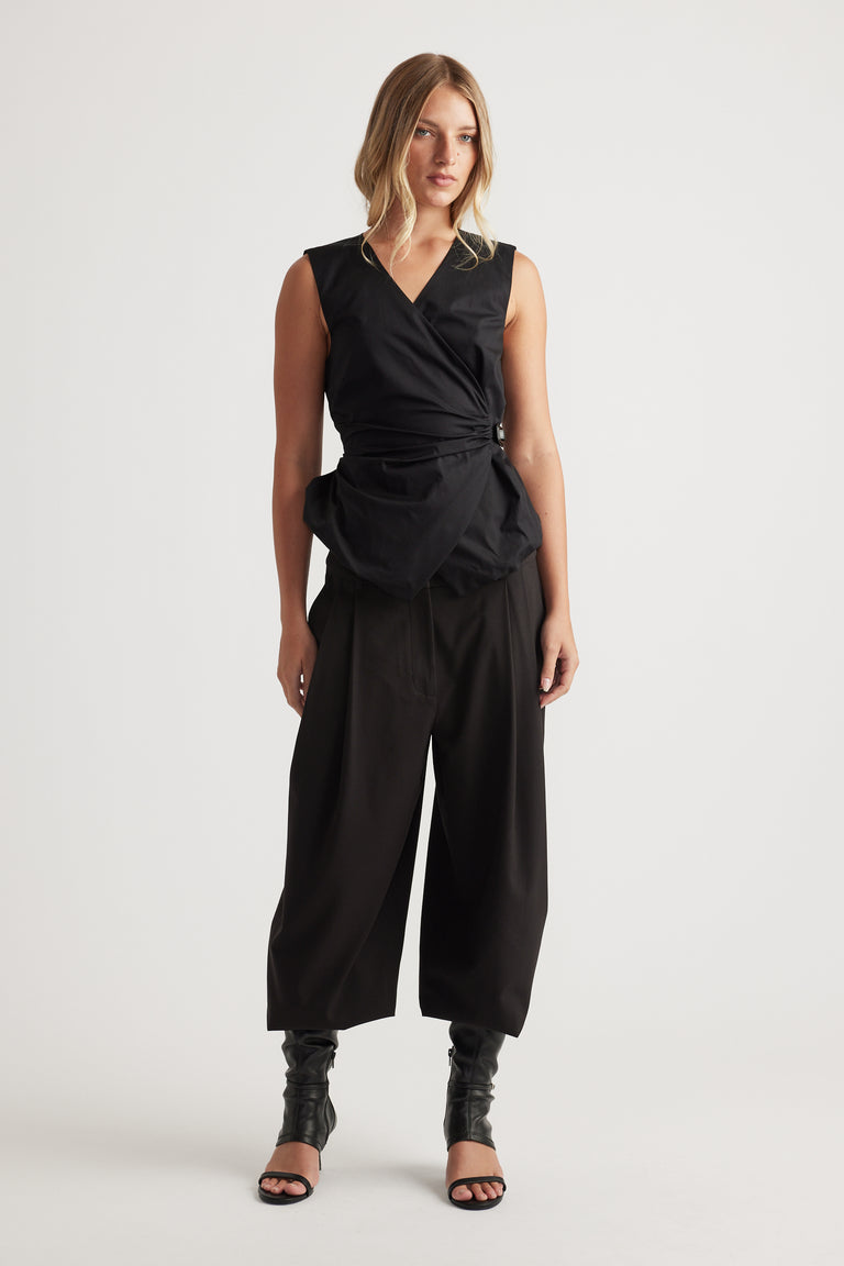 Woman wearing Ava Belted Wrap Top in black with sleeveless cut and adjustable side belt, styled with wide-leg pants by DECJUBA