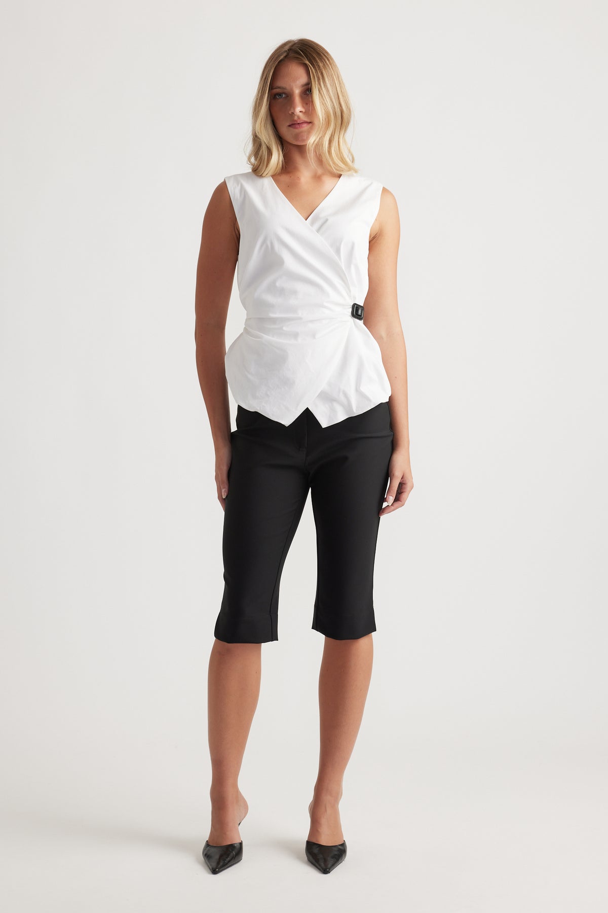 Woman wearing a sleeveless white wrap top with a side buckle paired with fitted black cropped pants and black heels