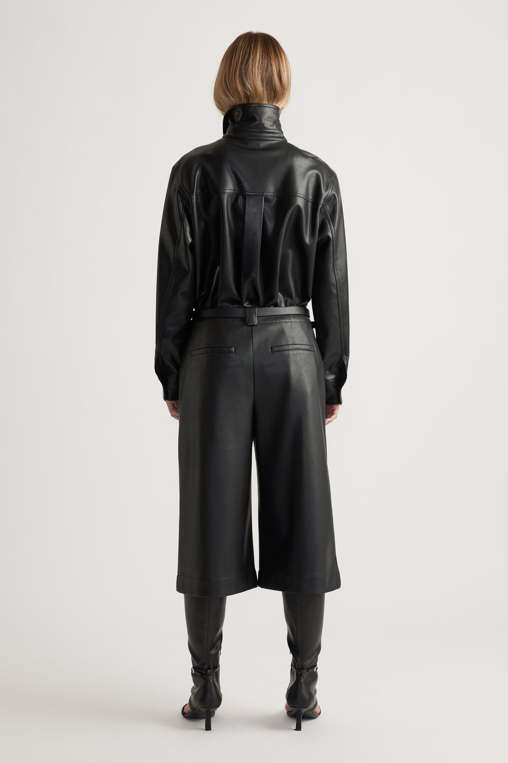 Woman shown from behind wearing Tabitha Faux Leather Short in black, styled with coordinating long-sleeve leather top and boots by DECJUBA