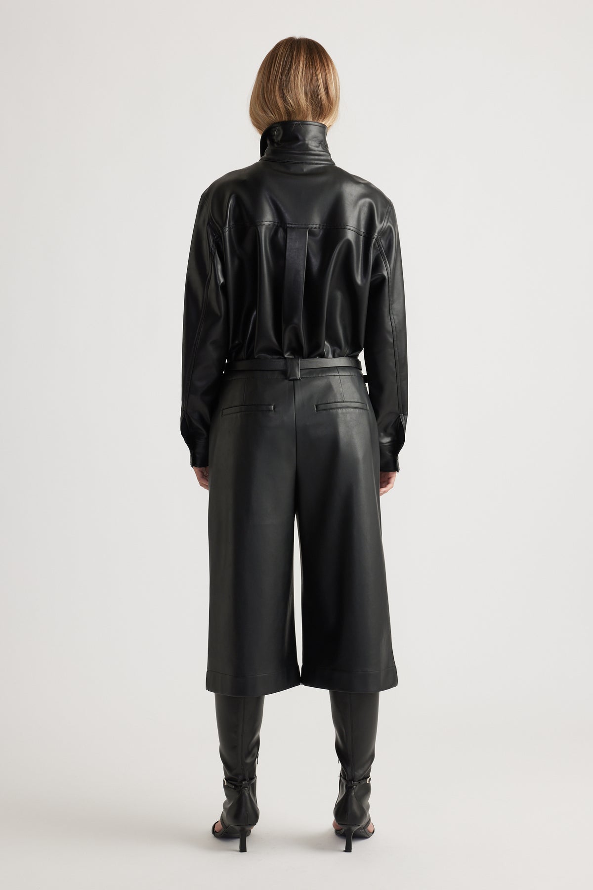 Woman shown from behind wearing Tabitha Faux Leather Short in black, styled with coordinating long-sleeve leather top and boots by DECJUBA