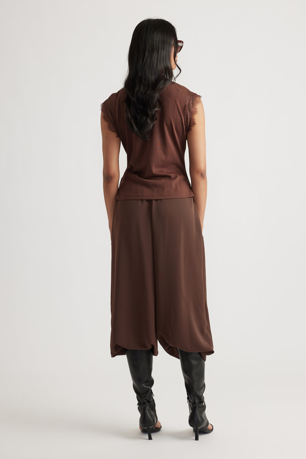 Woman with long dark hair wearing a sleeveless brown top and matching wide-leg pants with black boots, facing away