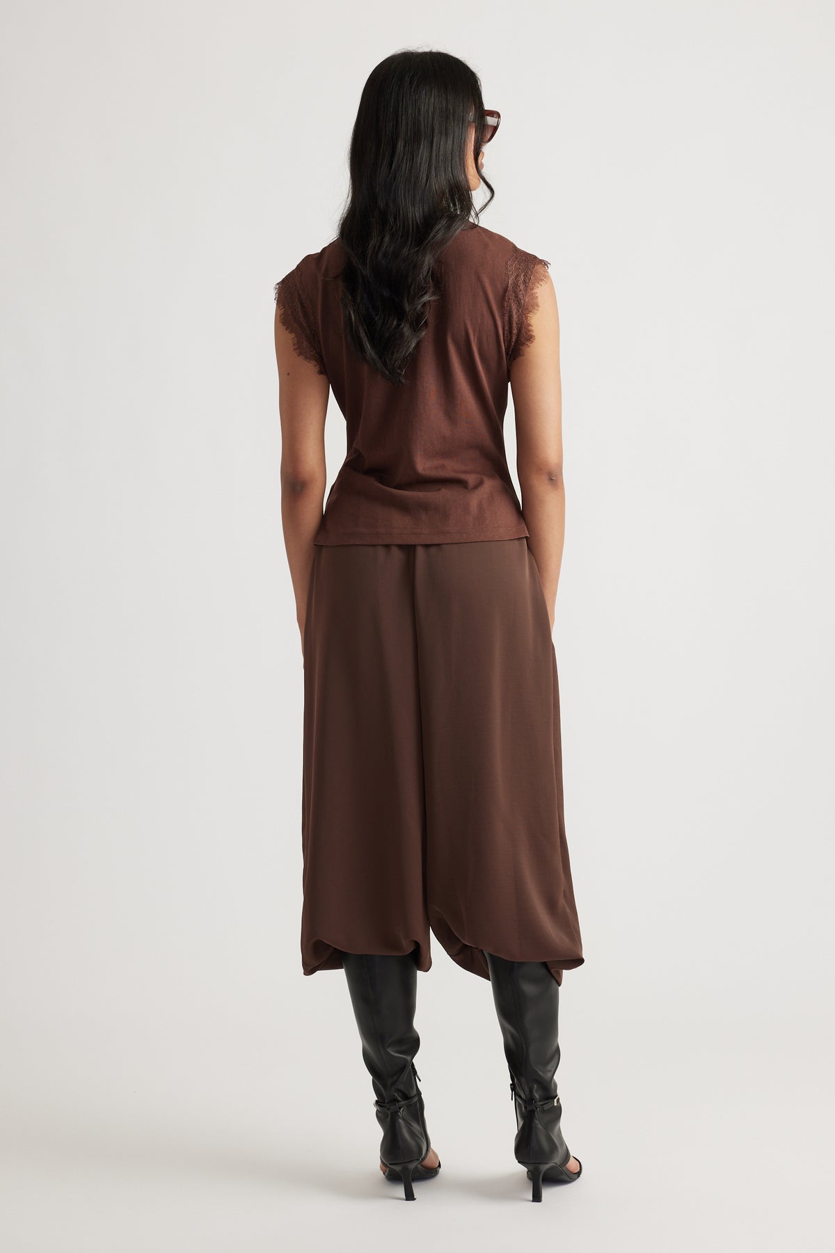 Woman with long dark hair wearing a sleeveless brown top and matching wide-leg pants with black boots, facing away