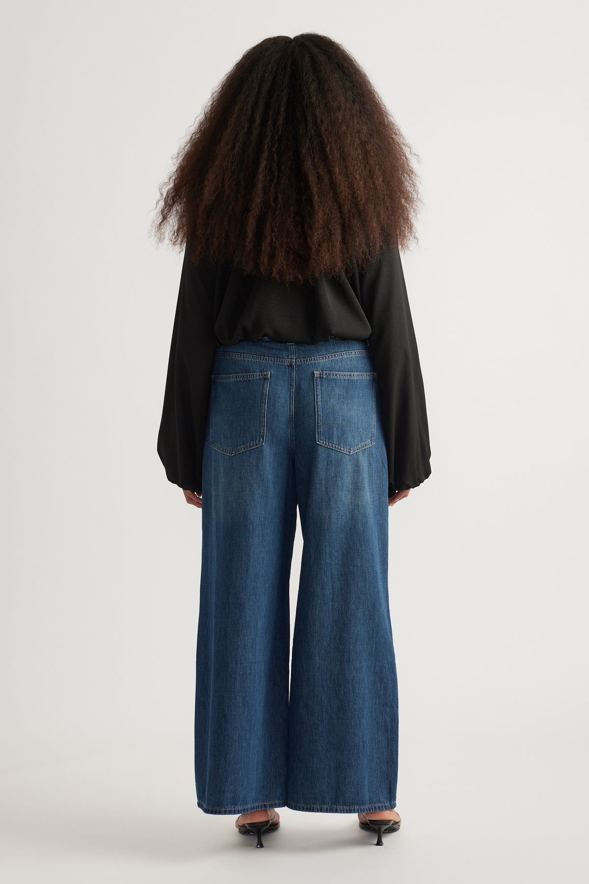 Back view of woman wearing full-length blue wide leg jeans with back pockets, a black long sleeve top and heels