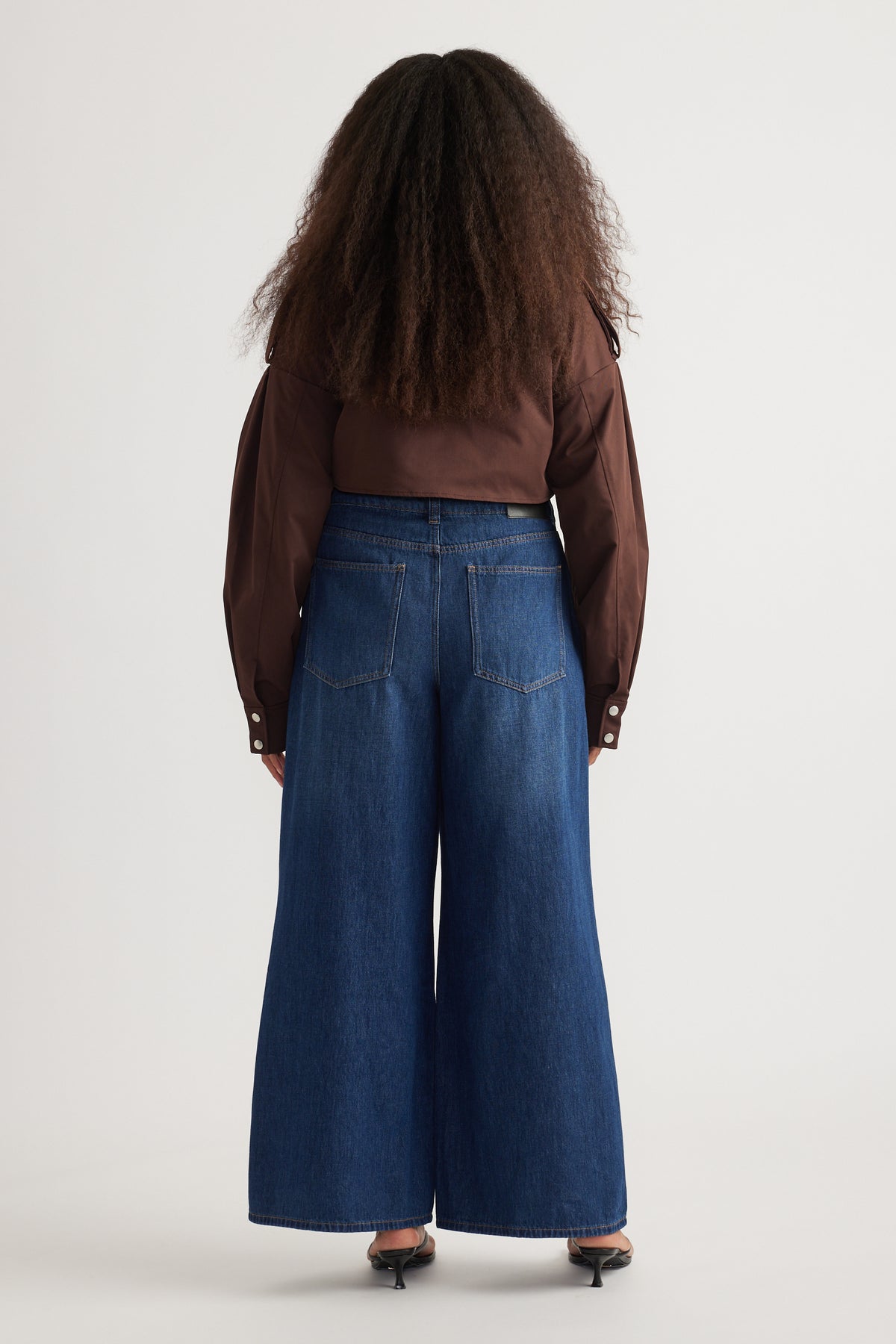 Back view of woman in dark blue full-length super wide-leg jeans, a brown shirt and heels