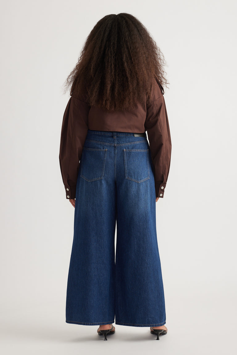 Back view of woman in dark blue full-length super wide-leg jeans, a brown shirt and heels