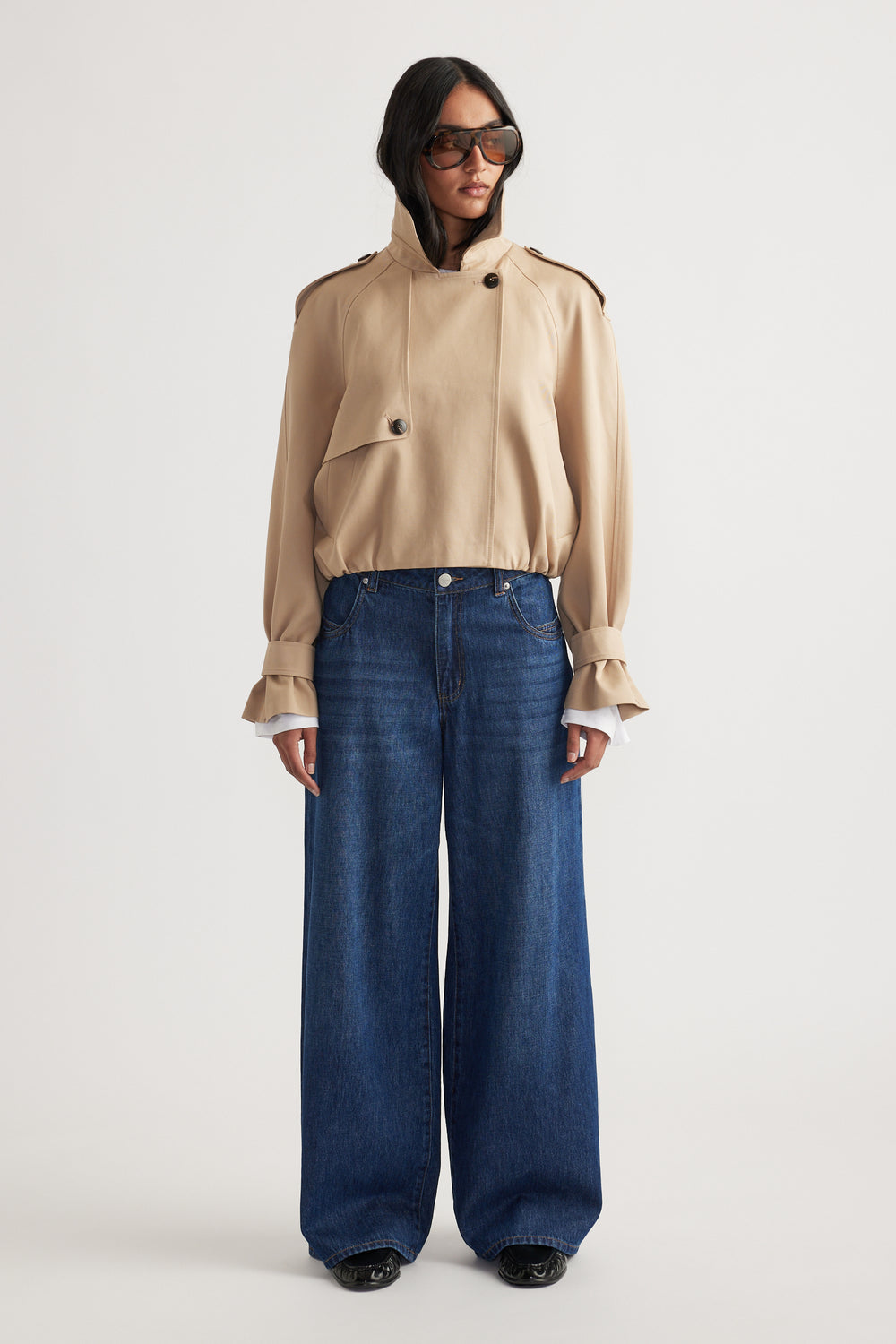 Woman wearing a beige high-neck jacket with button details paired with dark wide-leg denim jeans and oversized sunglasses