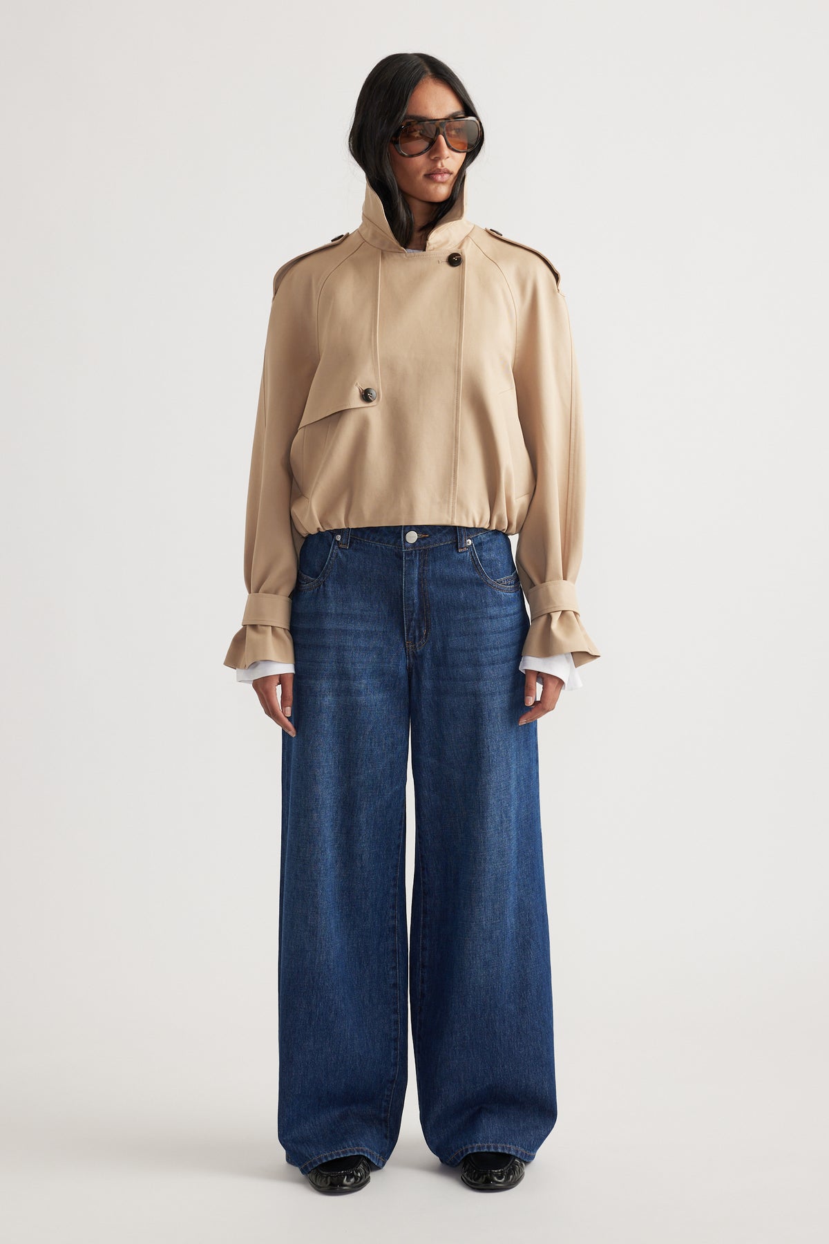 Woman wearing a beige high-neck jacket with button details paired with dark wide-leg denim jeans and oversized sunglasses