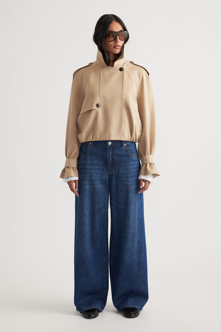 Woman wearing a beige high-neck jacket with button details paired with dark wide-leg denim jeans and oversized sunglasses