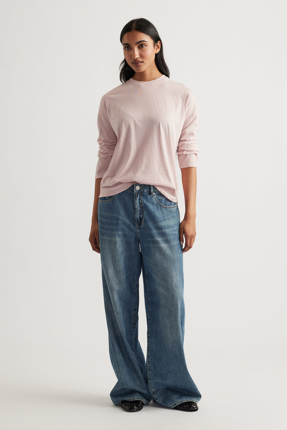 Woman with straight dark hair wearing a soft pink long sleeve top paired with relaxed wide-leg blue jeans facing forward