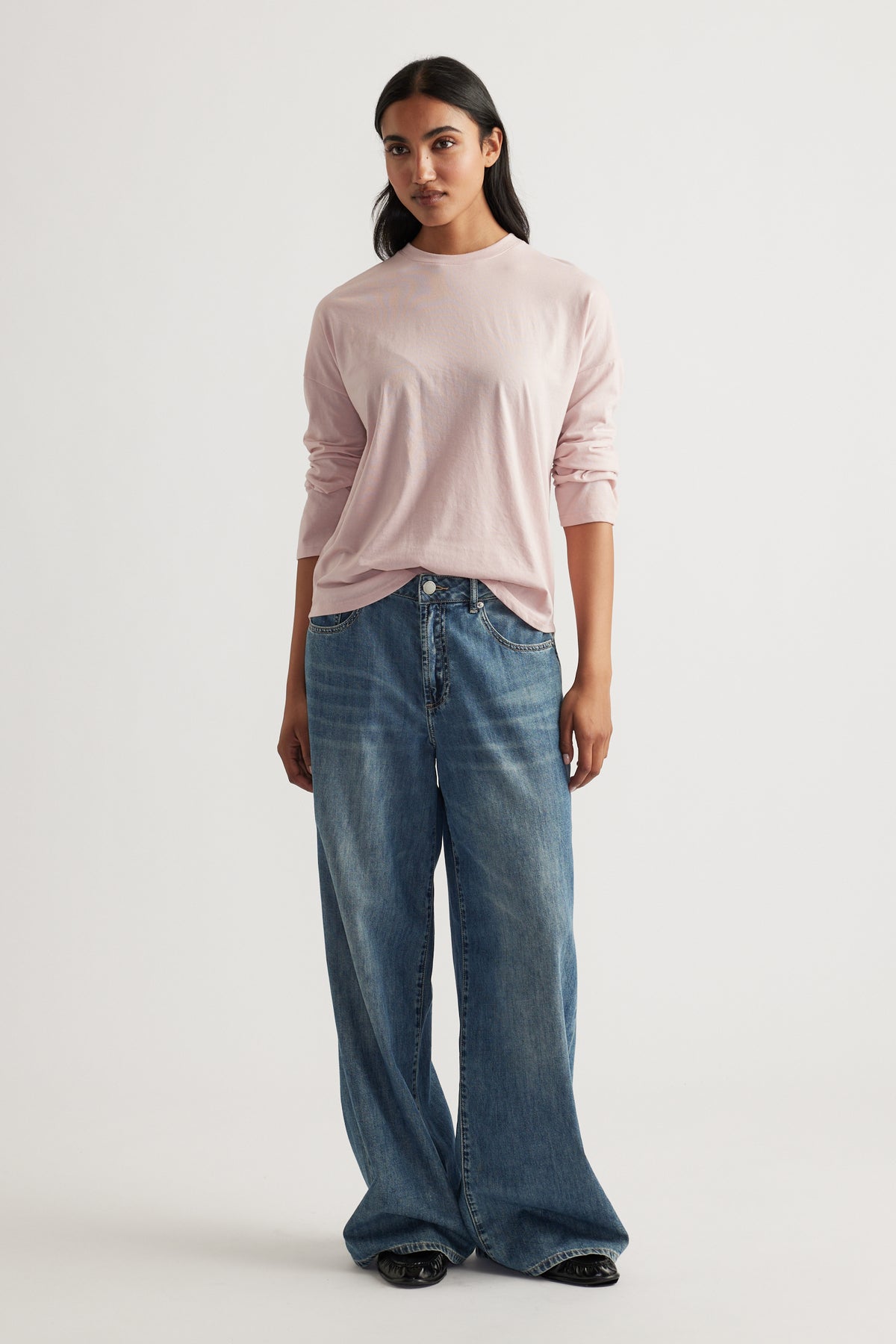 Woman with straight dark hair wearing a soft pink long sleeve top paired with relaxed wide-leg blue jeans facing forward