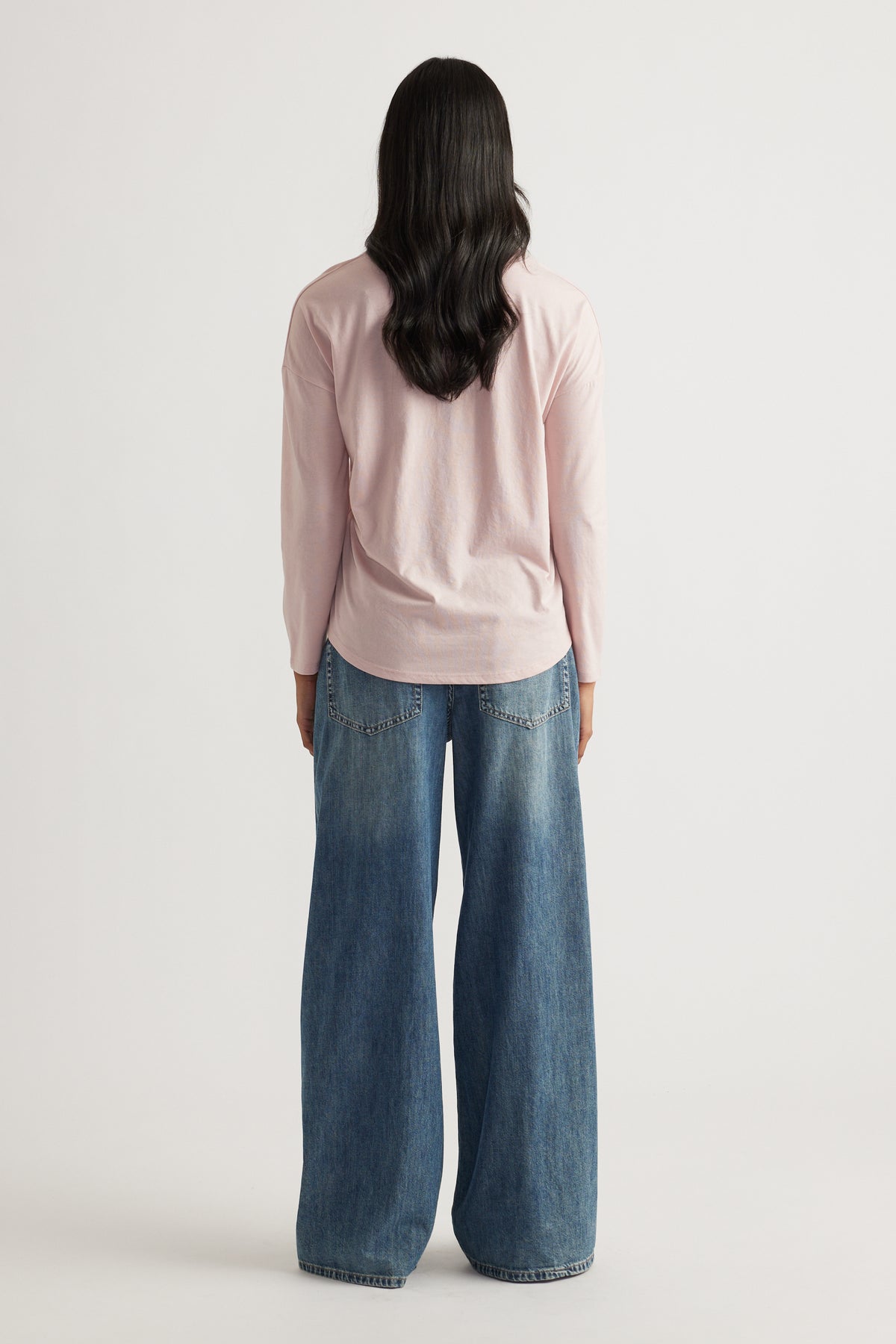 Person with long dark hair wearing a soft pink long sleeve top paired with wide-leg relaxed blue denim jeans viewed from behind