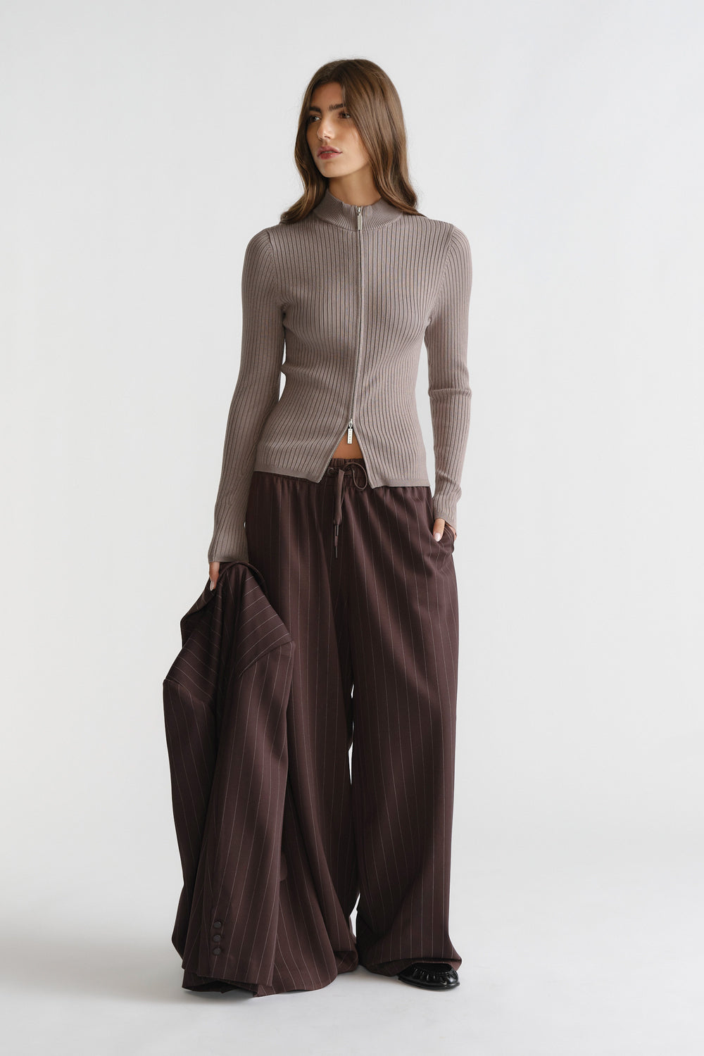 Woman wearing a taupe D-Luxe Knit Zip Long Sleeve paired with wide-leg pinstripe trousers and holding a matching jacket