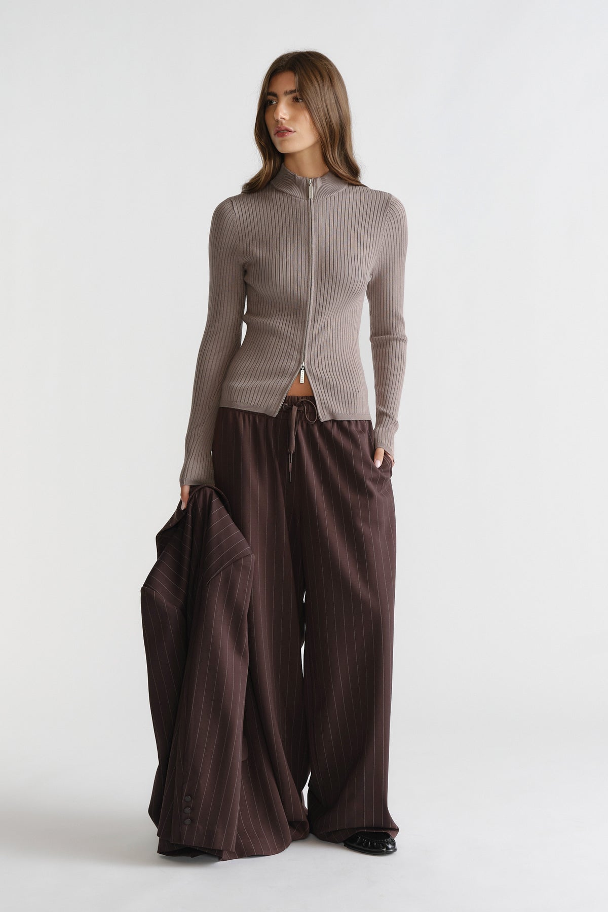 Woman wearing a taupe D-Luxe Knit Zip Long Sleeve paired with wide-leg pinstripe trousers and holding a matching jacket