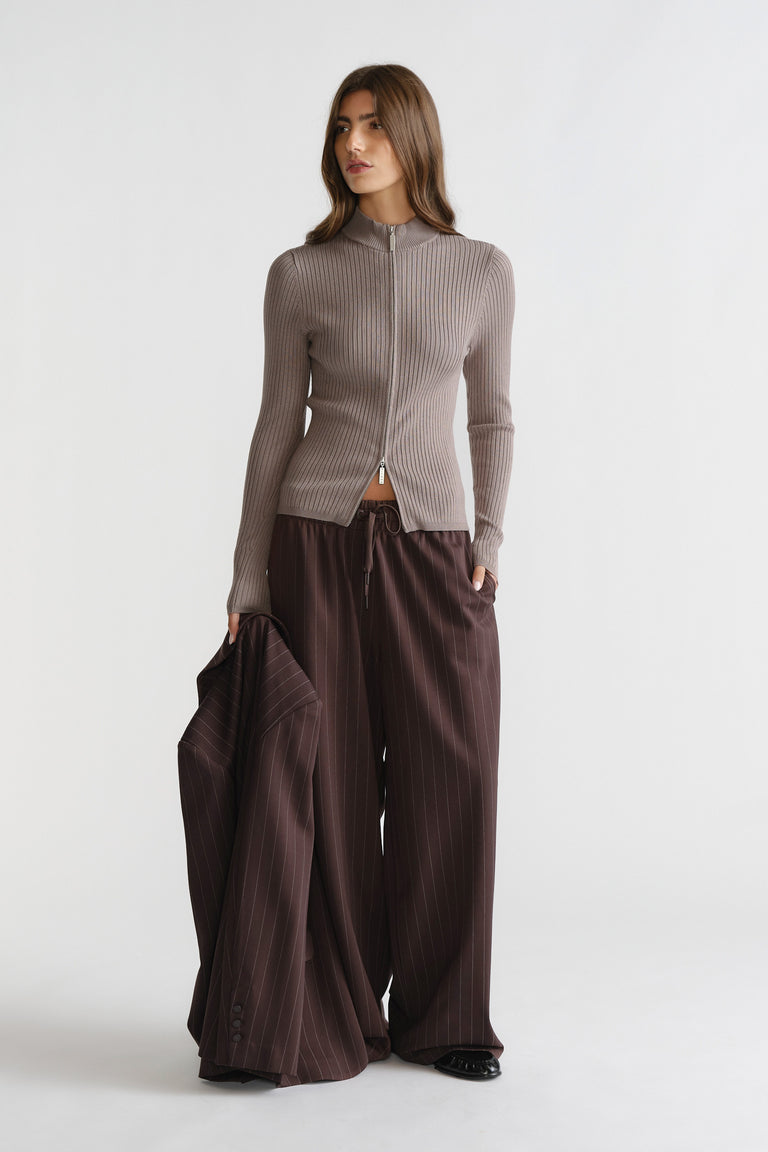 Woman wearing a taupe D-Luxe Knit Zip Long Sleeve paired with wide-leg pinstripe trousers and holding a matching jacket