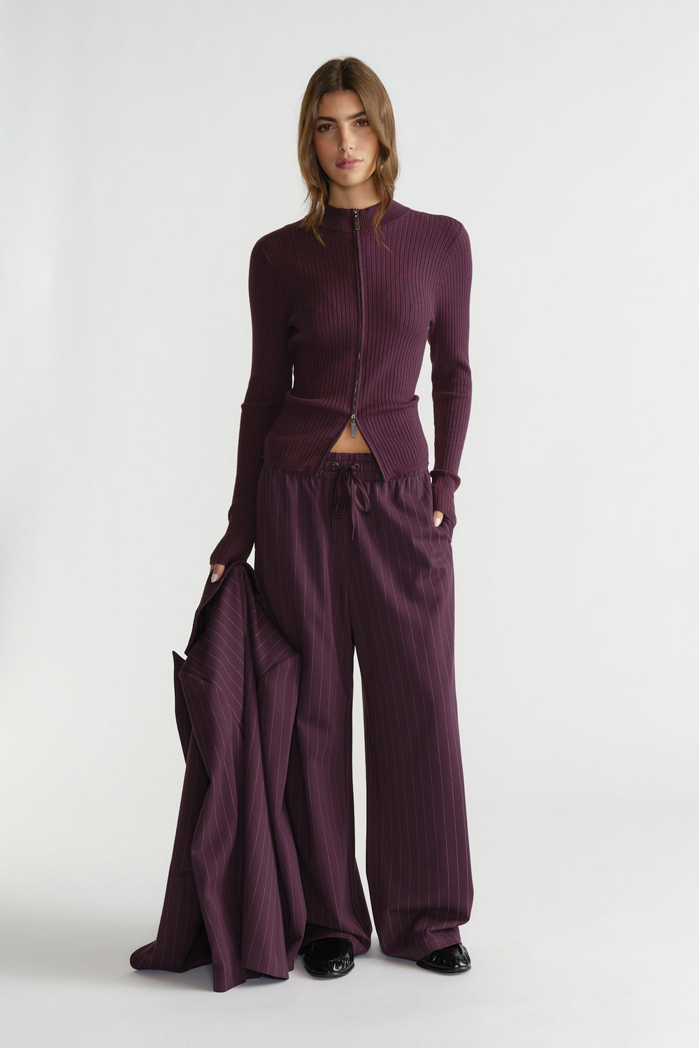 Woman wearing a fitted ribbed zip knit top and loose tailored pants in deep plum, holding a matching pinstripe blazer.
