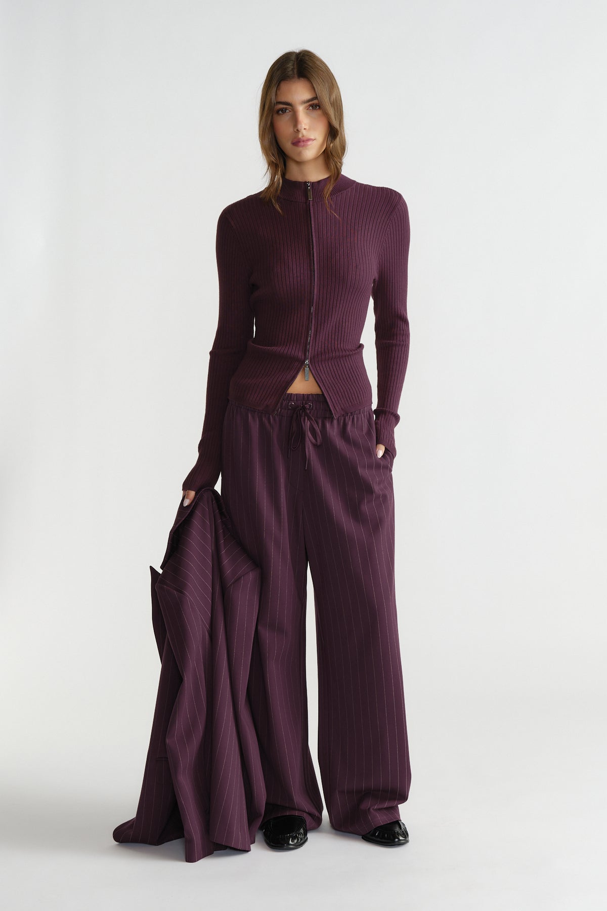 Woman wearing a fitted ribbed zip knit top and loose tailored pants in deep plum, holding a matching pinstripe blazer.