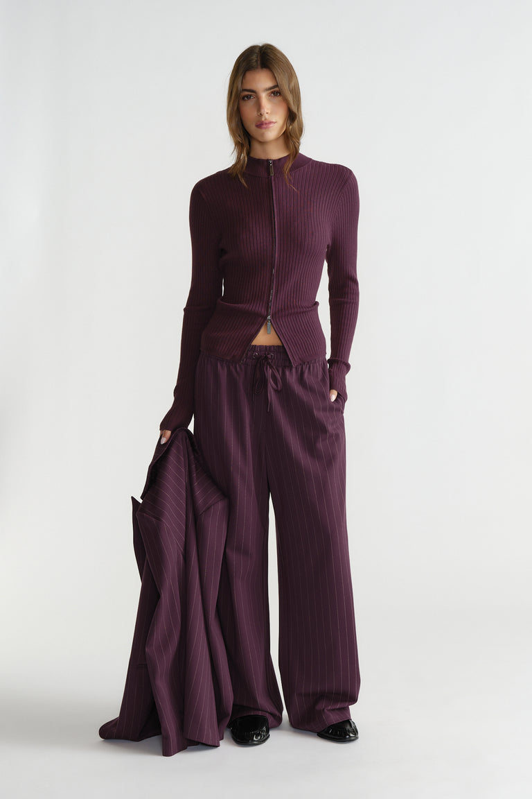 Woman wearing a fitted ribbed zip knit top and loose tailored pants in deep plum, holding a matching pinstripe blazer.