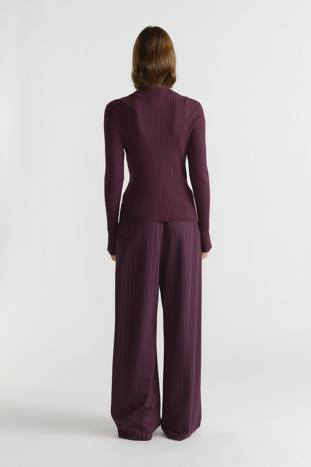 Back view of woman wearing a ribbed, long sleeve knit top paired with wide-leg pinstriped trousers in deep purple.