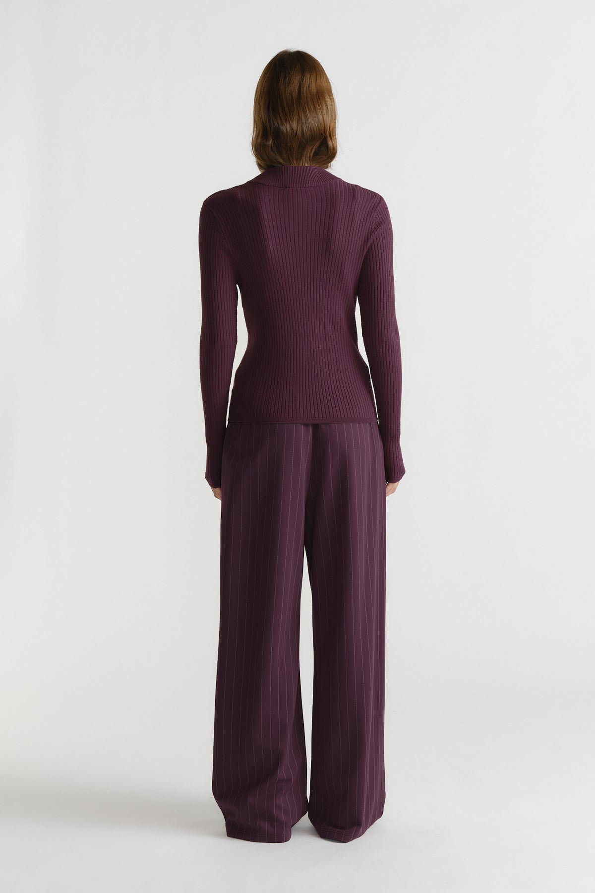 Back view of woman wearing a ribbed, long sleeve knit top paired with wide-leg pinstriped trousers in deep purple.