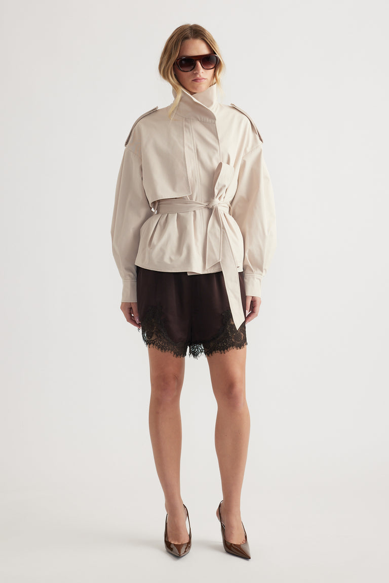 Woman wearing a belted light beige cropped trench with epaulettes paired with black lace-trimmed shorts and brown heels