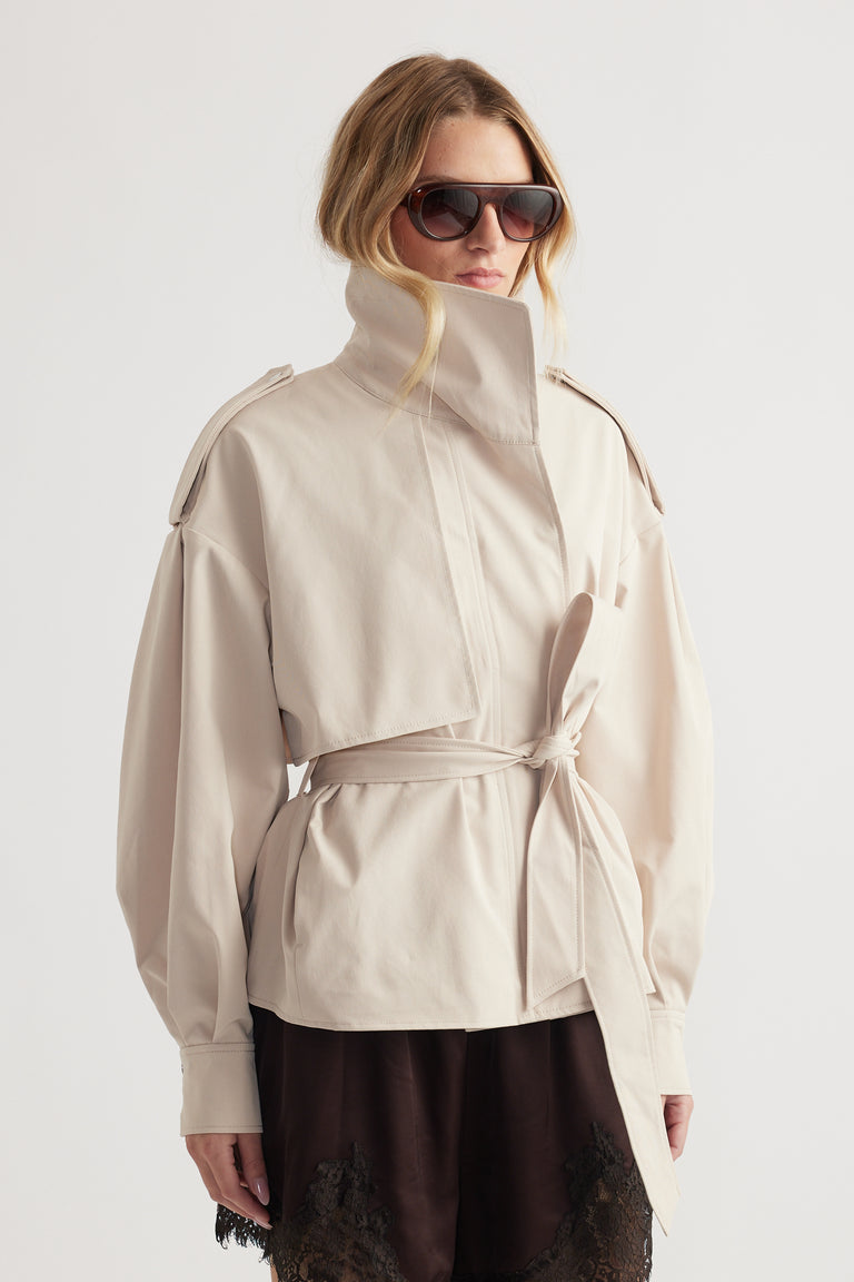 Woman wearing a beige cropped trench with high collar and belted waist paired with dark lace-trimmed shorts and sunglasses