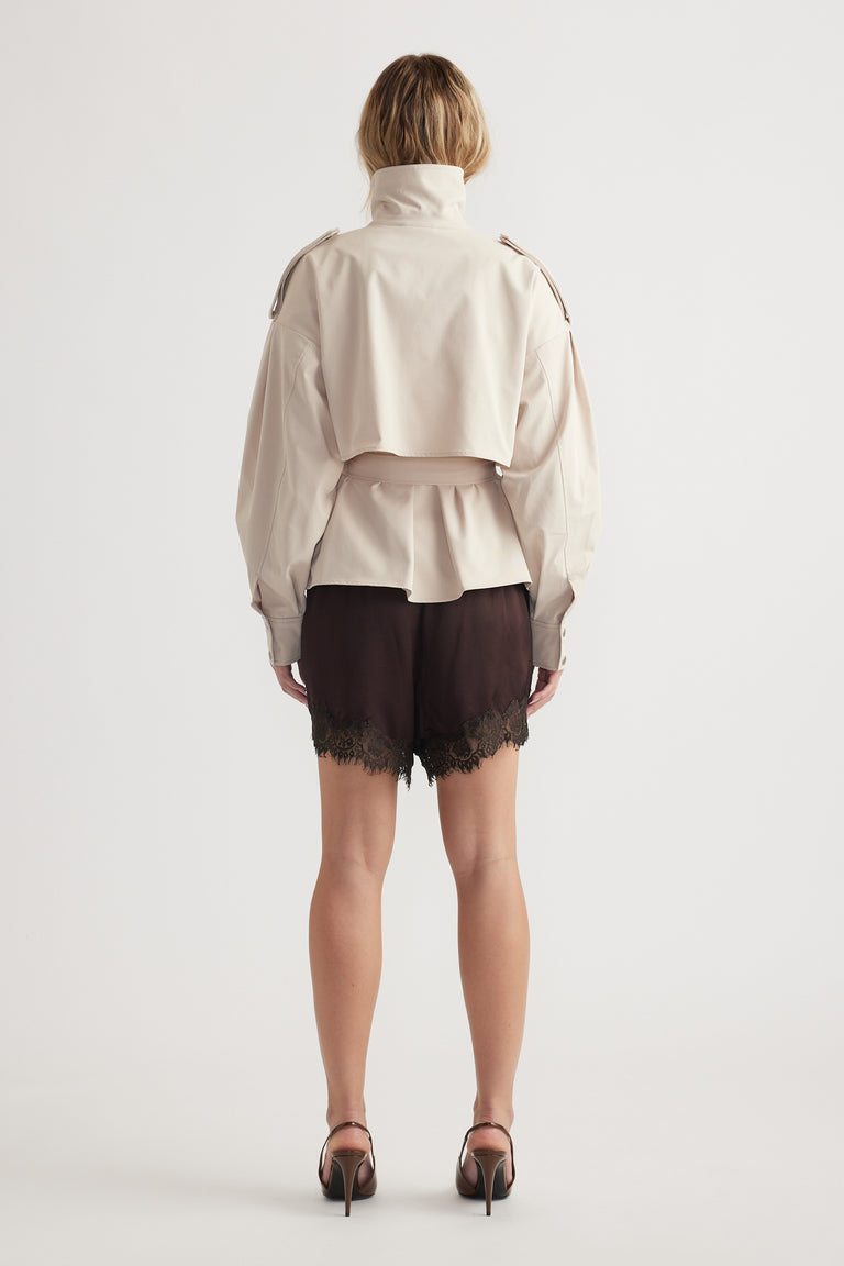 Woman wearing a cropped beige trench with epaulettes paired with dark lace-trimmed shorts and black heels viewed from behind