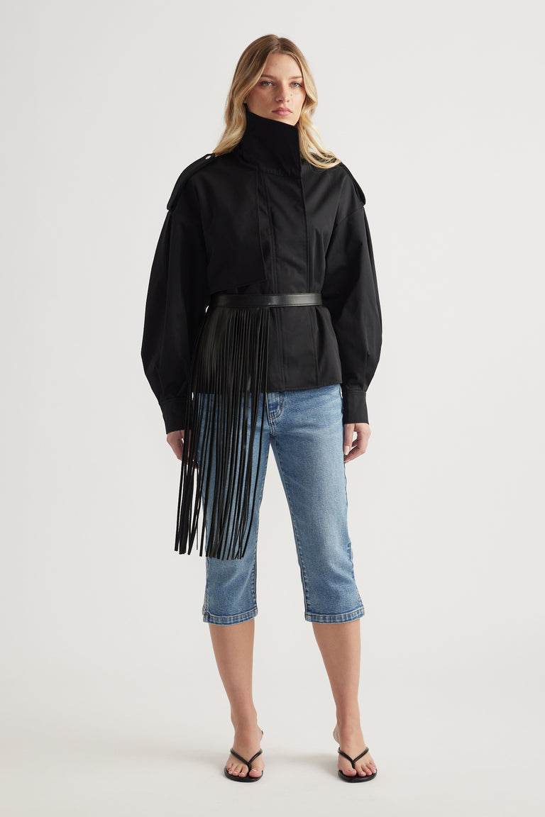 Woman wearing black structured jacket with high collar and fringed belt paired with cropped blue denim jeans and sandals