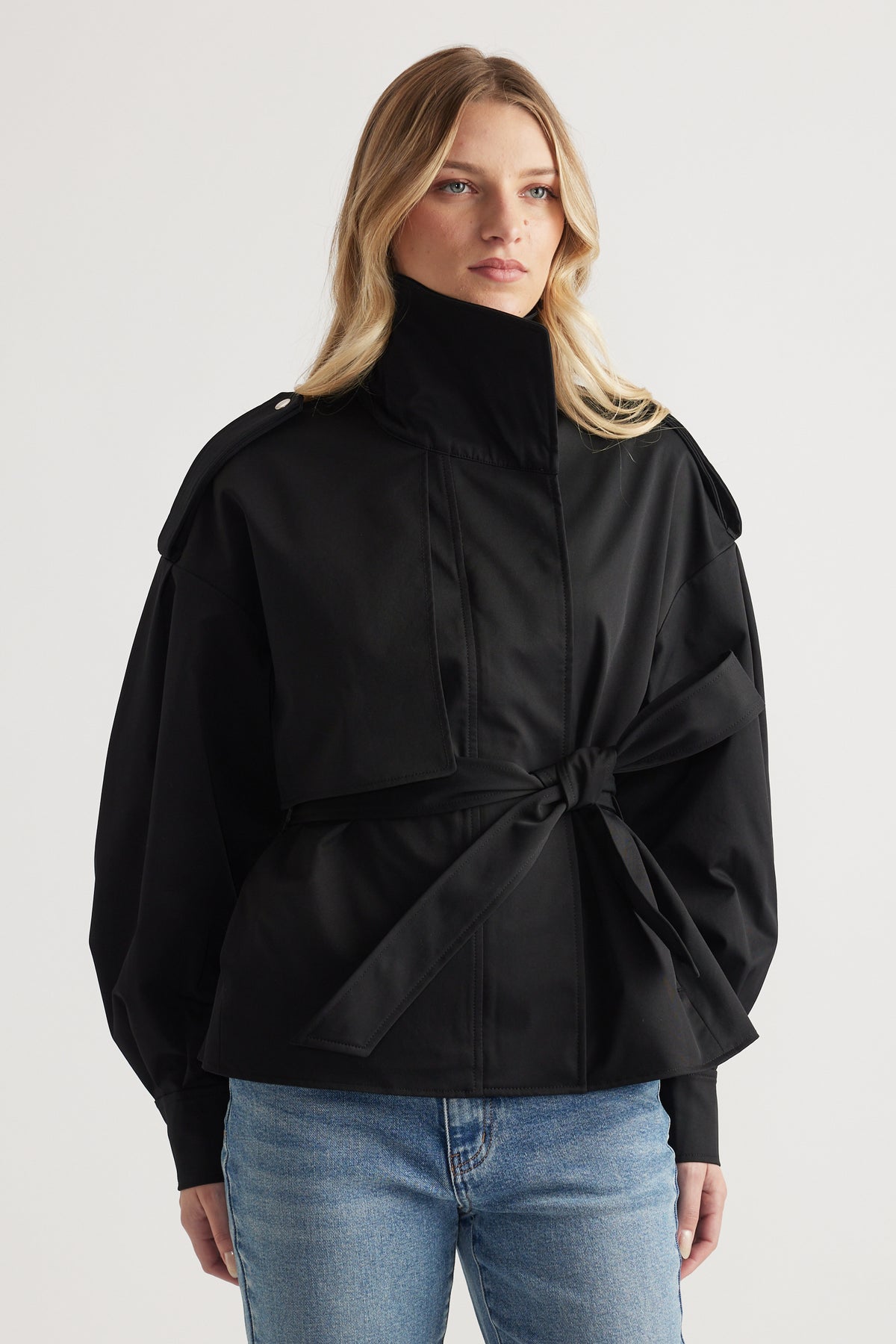 Woman wearing a belted black cropped trench coat with structured shoulders and high collar paired with blue jeans