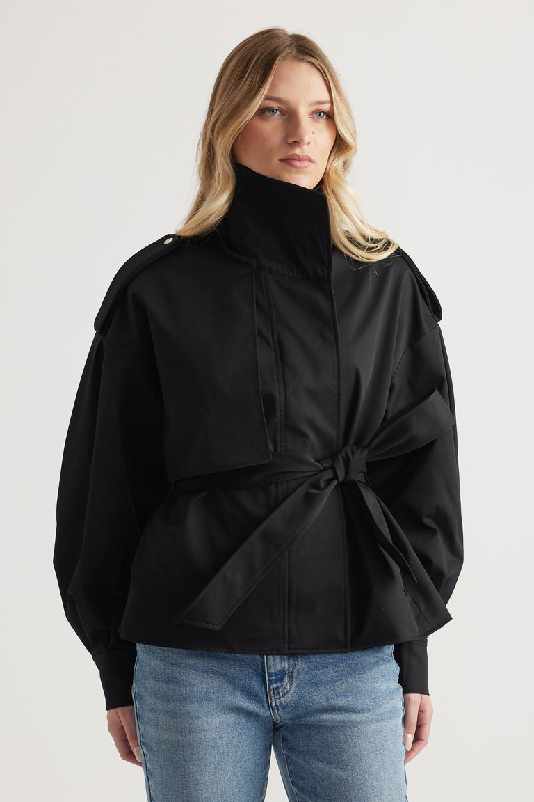 Woman wearing a belted black cropped trench coat with structured shoulders and high collar paired with blue jeans