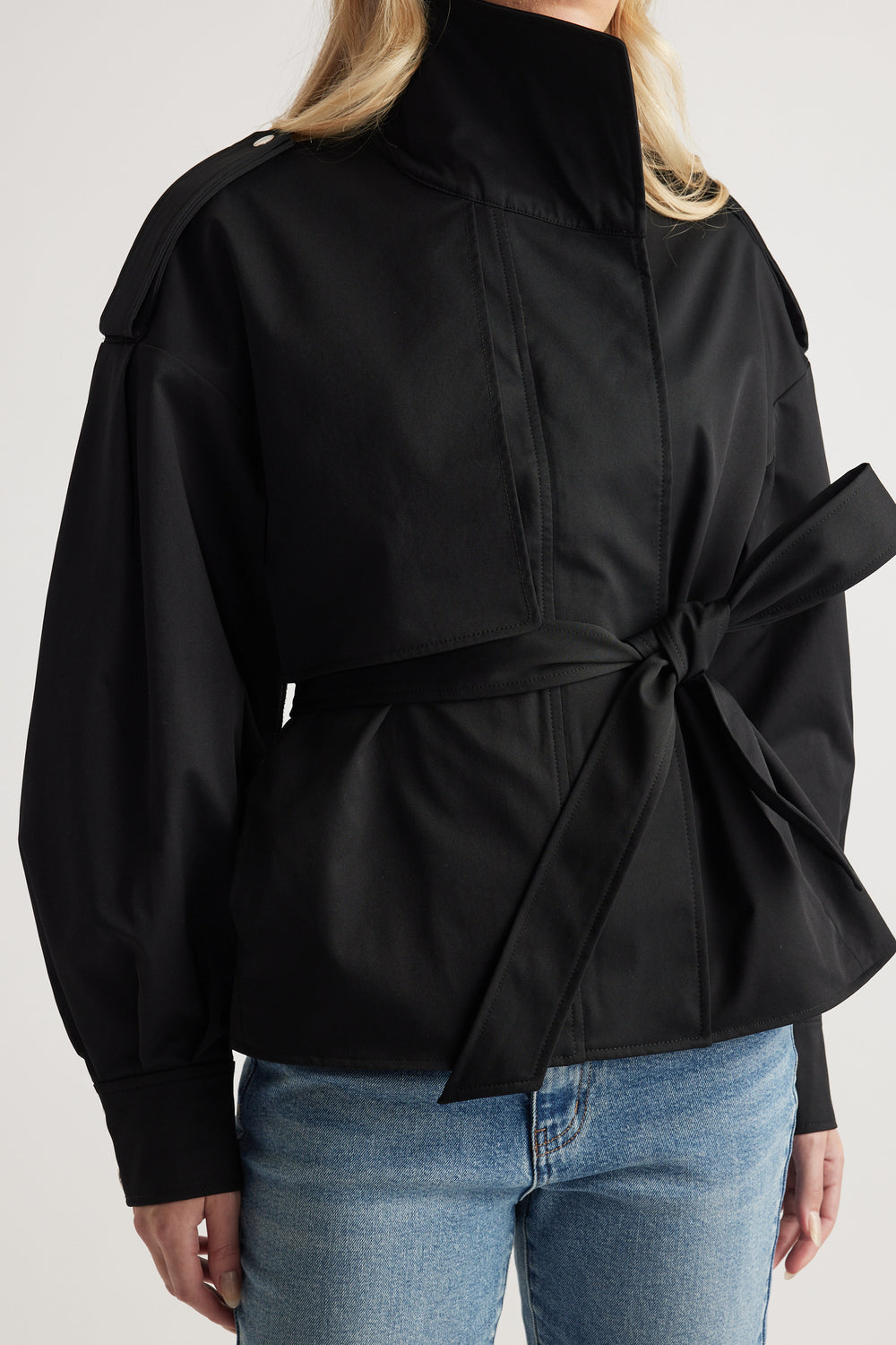 Woman wearing black cropped trench with high collar, shoulder epaulettes, tied waist belt, paired with light blue jeans