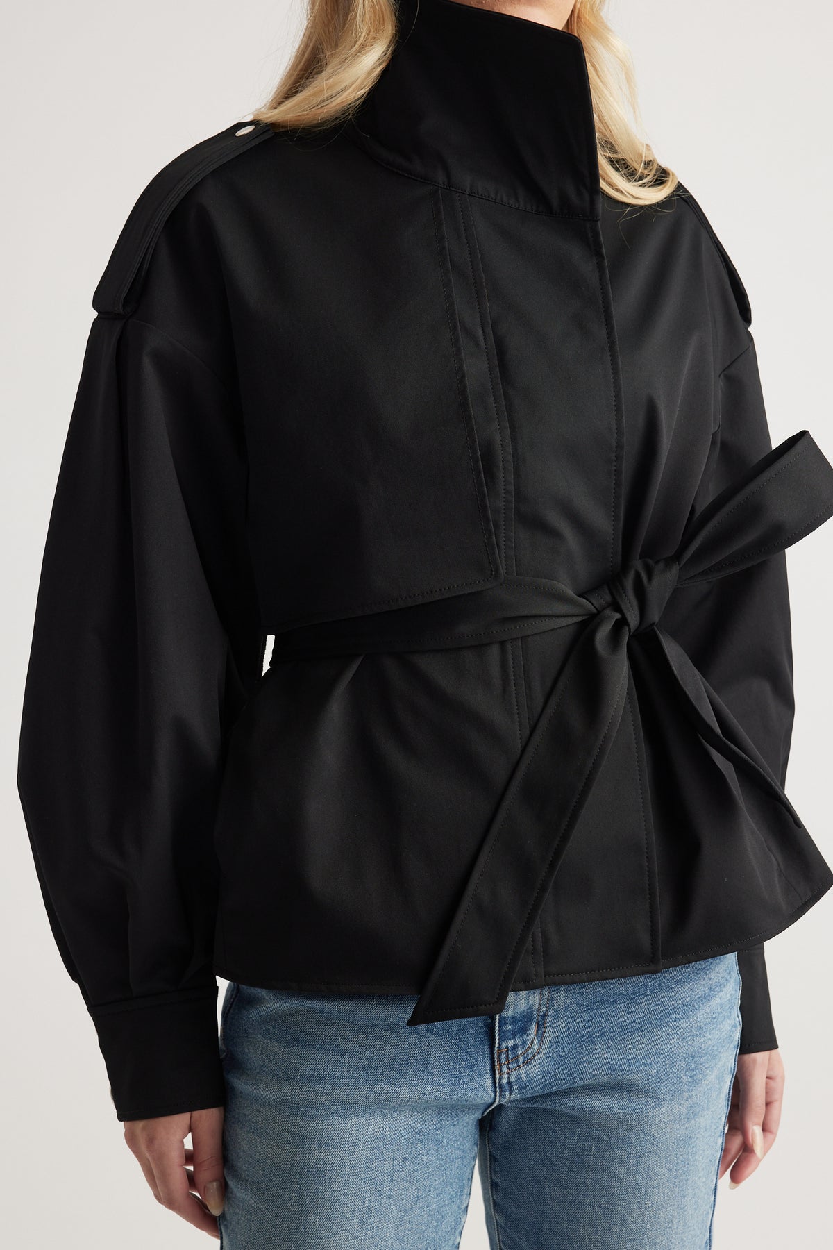 Woman wearing black cropped trench with high collar, shoulder epaulettes, tied waist belt, paired with light blue jeans
