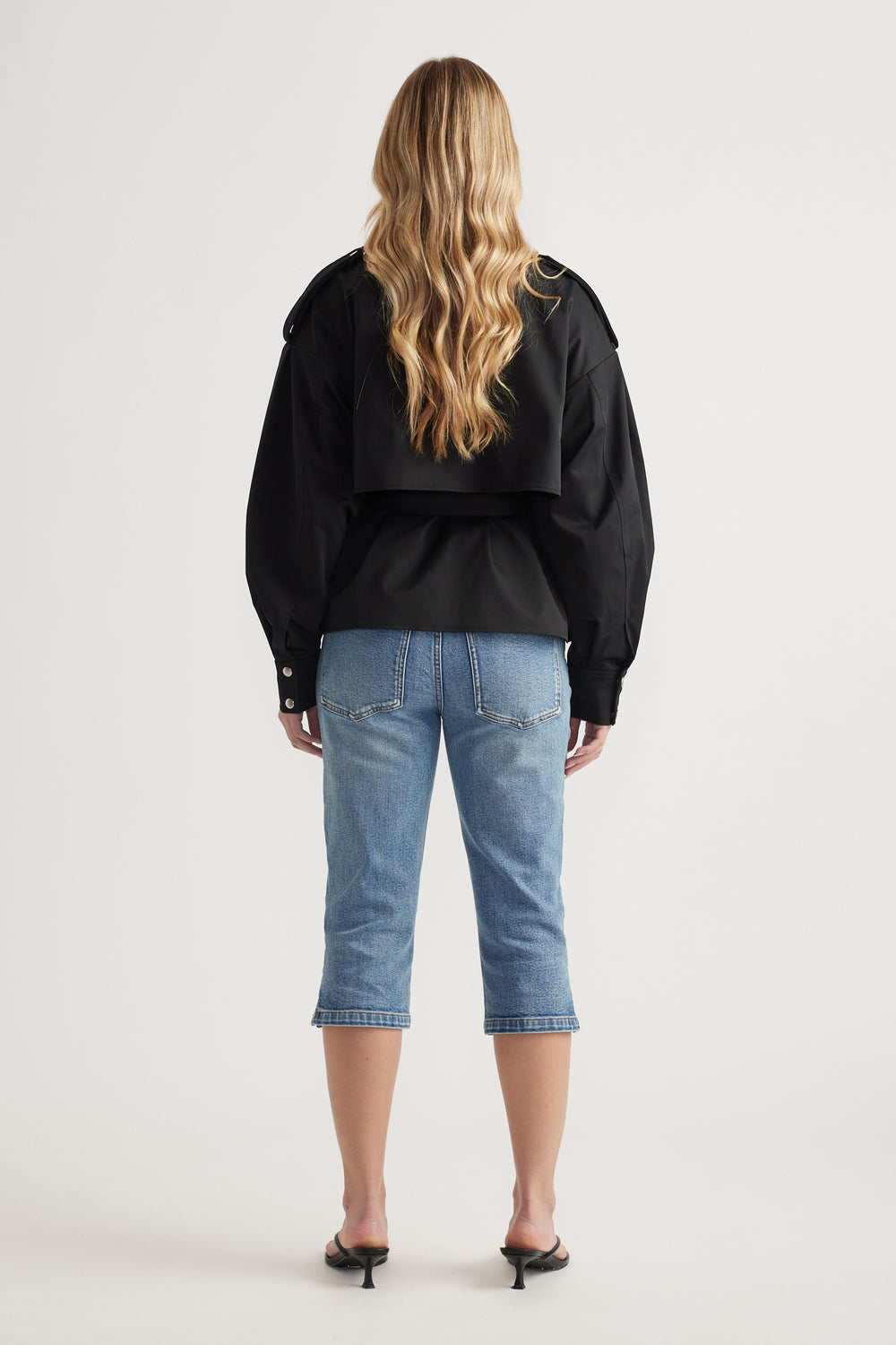 Woman with long wavy blonde hair wearing a black cropped jacket with epaulettes and mid-blue cropped denim jeans