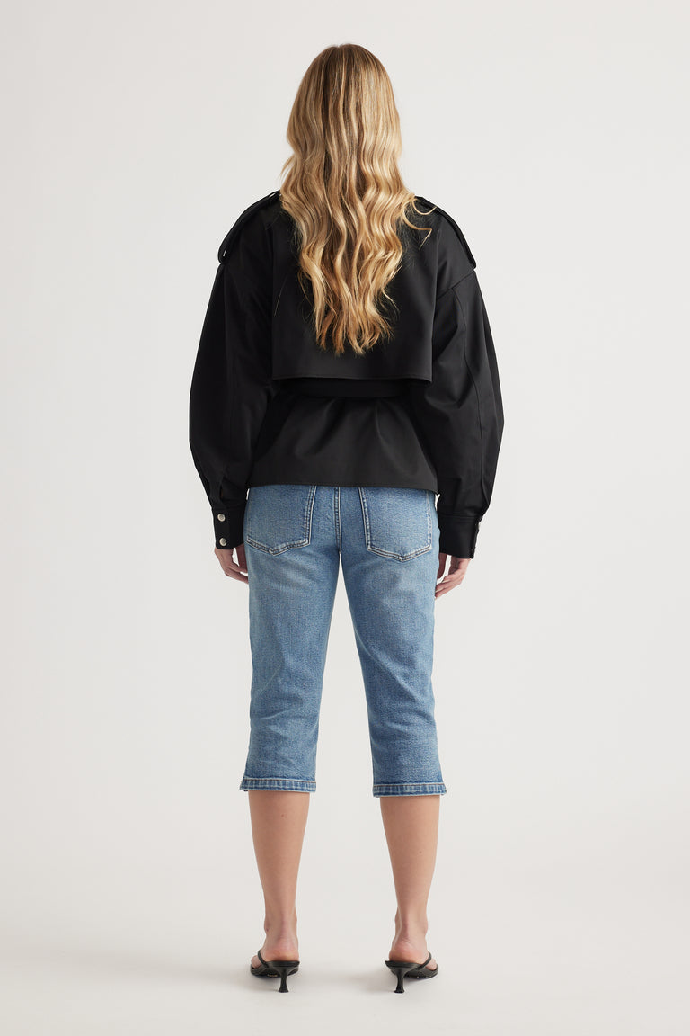 Woman with long wavy blonde hair wearing a black cropped jacket with epaulettes and mid-blue cropped denim jeans